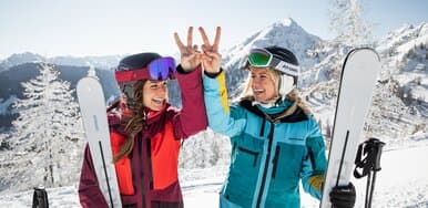 Two female skiers stand laughing on the ski slope, holding Atomic skis and making a peace sign with their hands, symbolizing the 4-mountain ski area Schladming, with snowy mountain scenery in the background. | © Mirja Geh